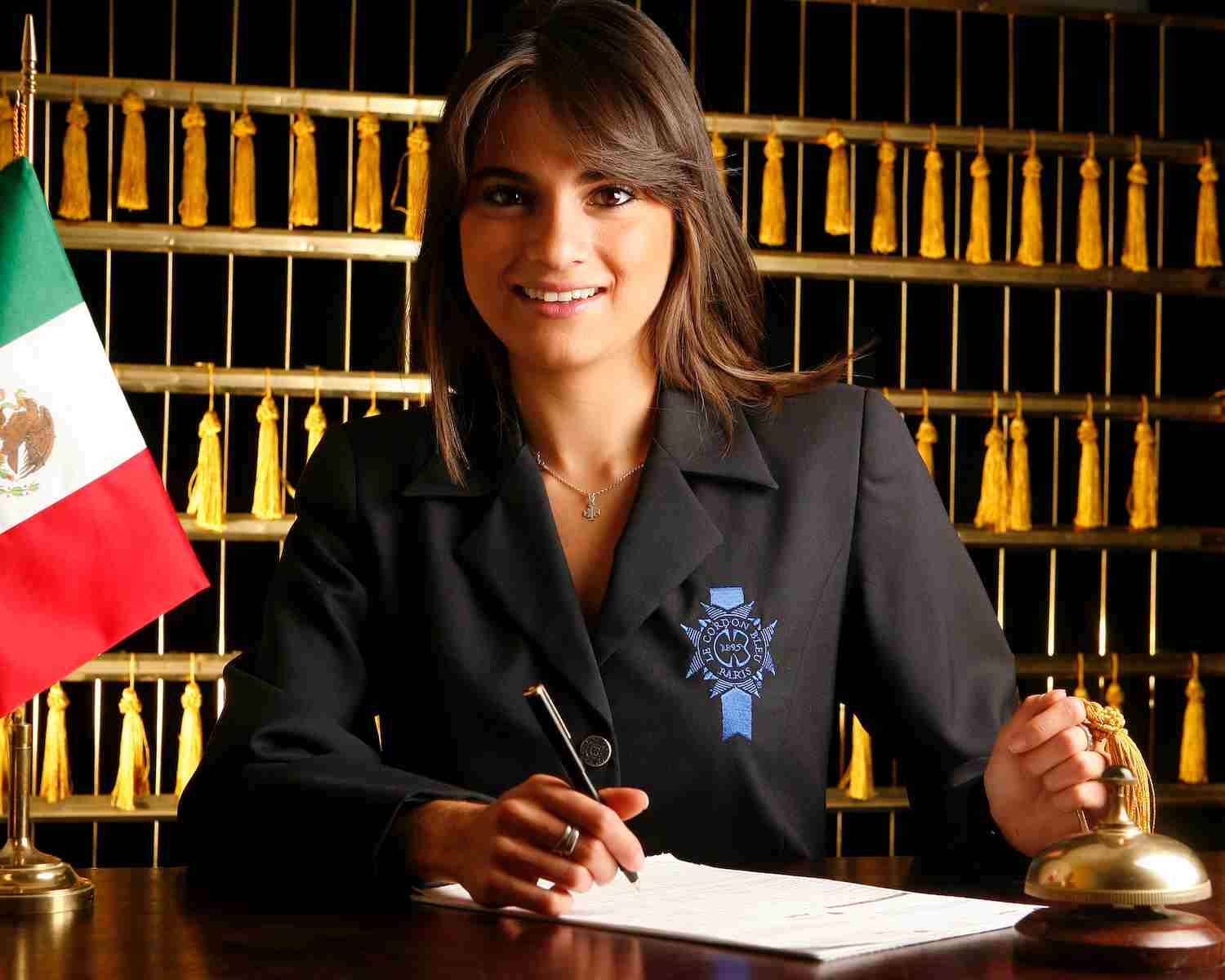 Mujer sonriente con uniforme formal escribe en un registro junto a una campana de hotel, con una bandera de México a un lado y un fondo de llaves doradas colgadas.